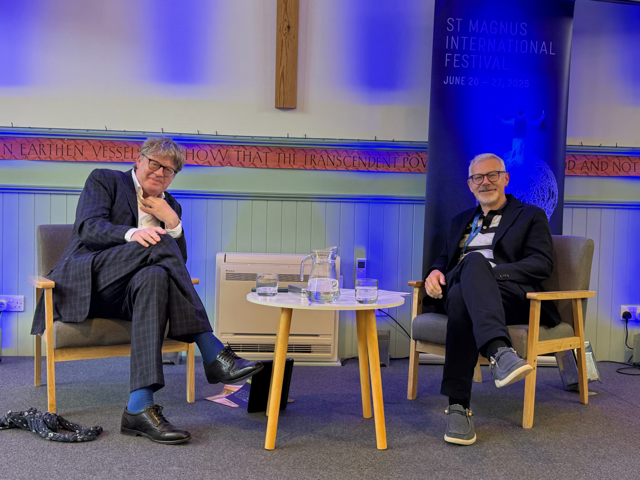 James Runcie seated on stage in conversation with Alasdair Nicolson beneath a St Magnus International Festival banner